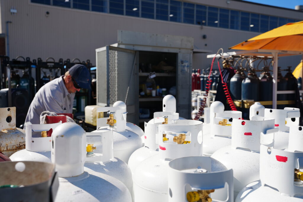 Rows of white portable propane tanks at an outdoor refill yard, with a worker and other gas cylinders in the background