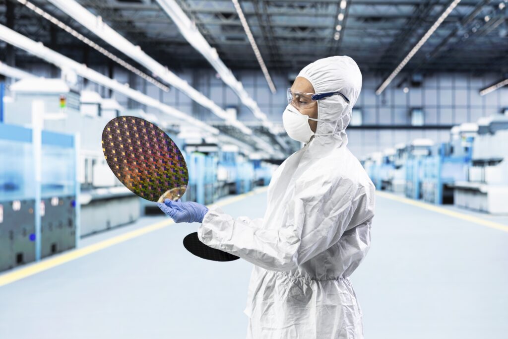 Technician in a cleanroom suit inspecting a patterned silicon wafer on a semiconductor fab floor