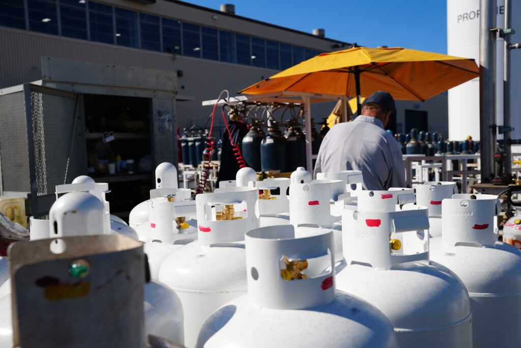 Rows of propane cylinders stored outdoors, with a worker filling cylinders in the background