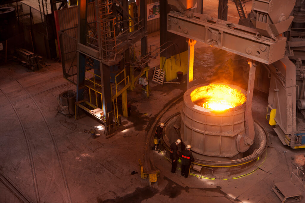 Three workers in hard hats stand beside a container of glowing molten metal inside a steel mill