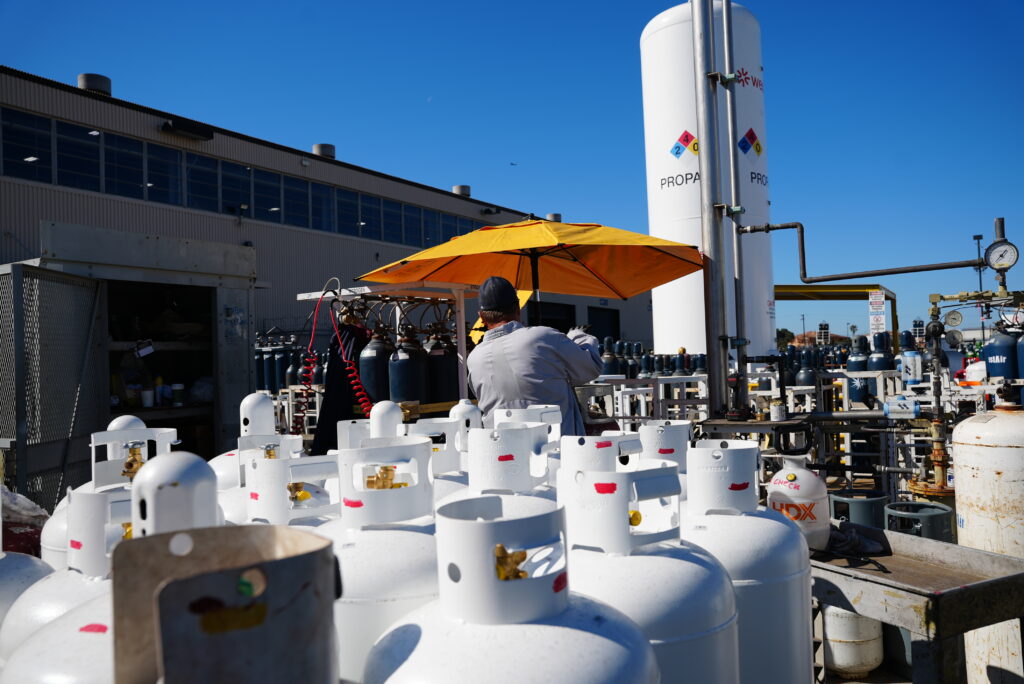 Rows of white propane cylinders stored outdoors at ground level in a WestAir fill yard, with a bulk propane tank and a secured cylinder cage in the background